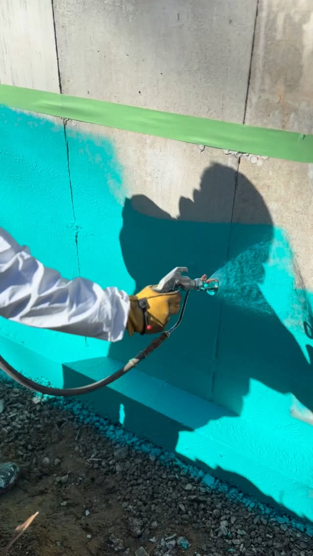Crew member spray-applying a teal waterproofing membrane to a foundation wall